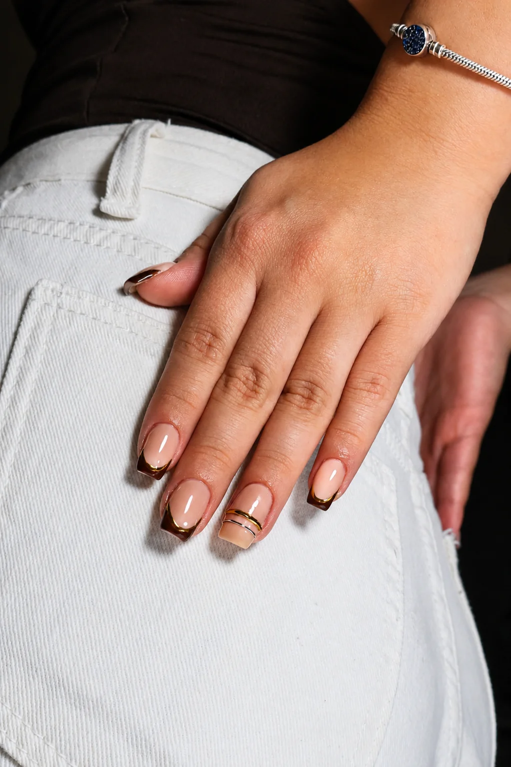 Mano apoyada sobre tela blanca con manicura francesa en marrón y línea dorada.